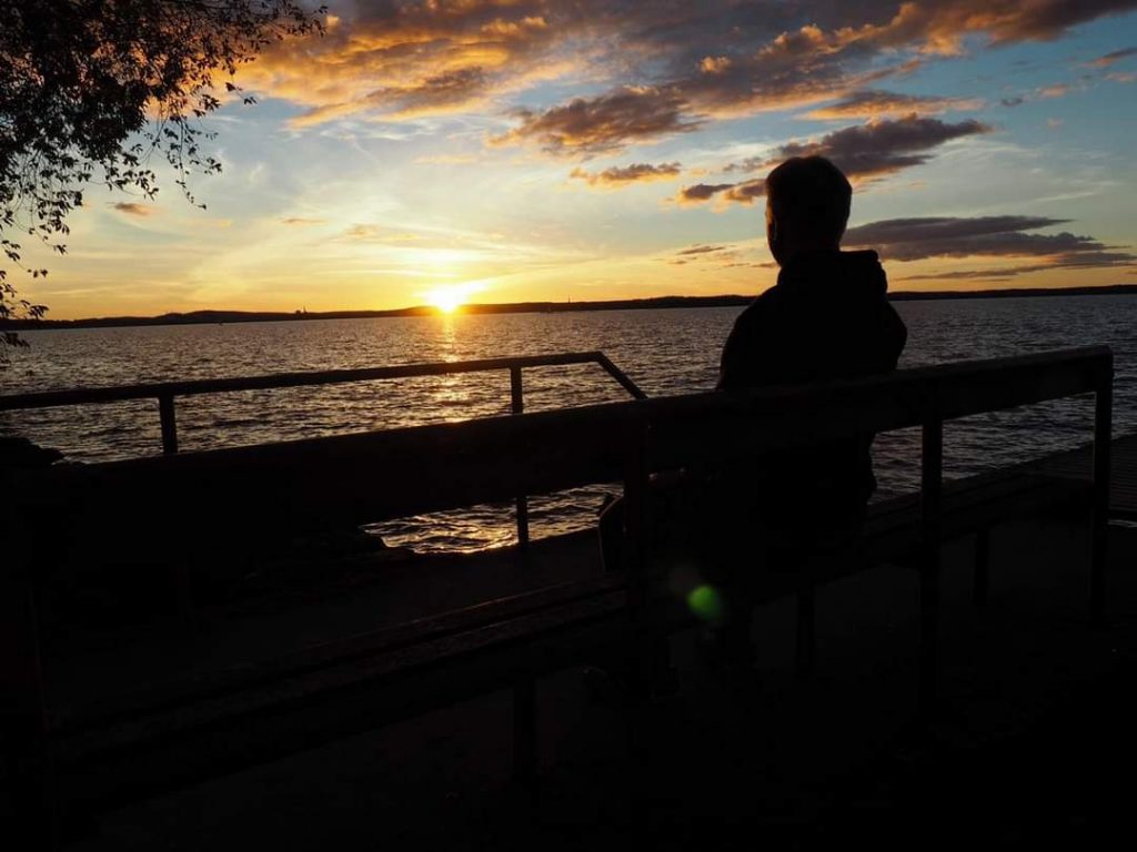 Mies istuu laiturilla ja katsoo, kun aurinko laskeutuu järven taakse.
